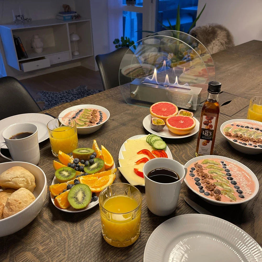 Circular table bioethanol fireplace lit on a dining table set with breakfast plates, fruit and coffee